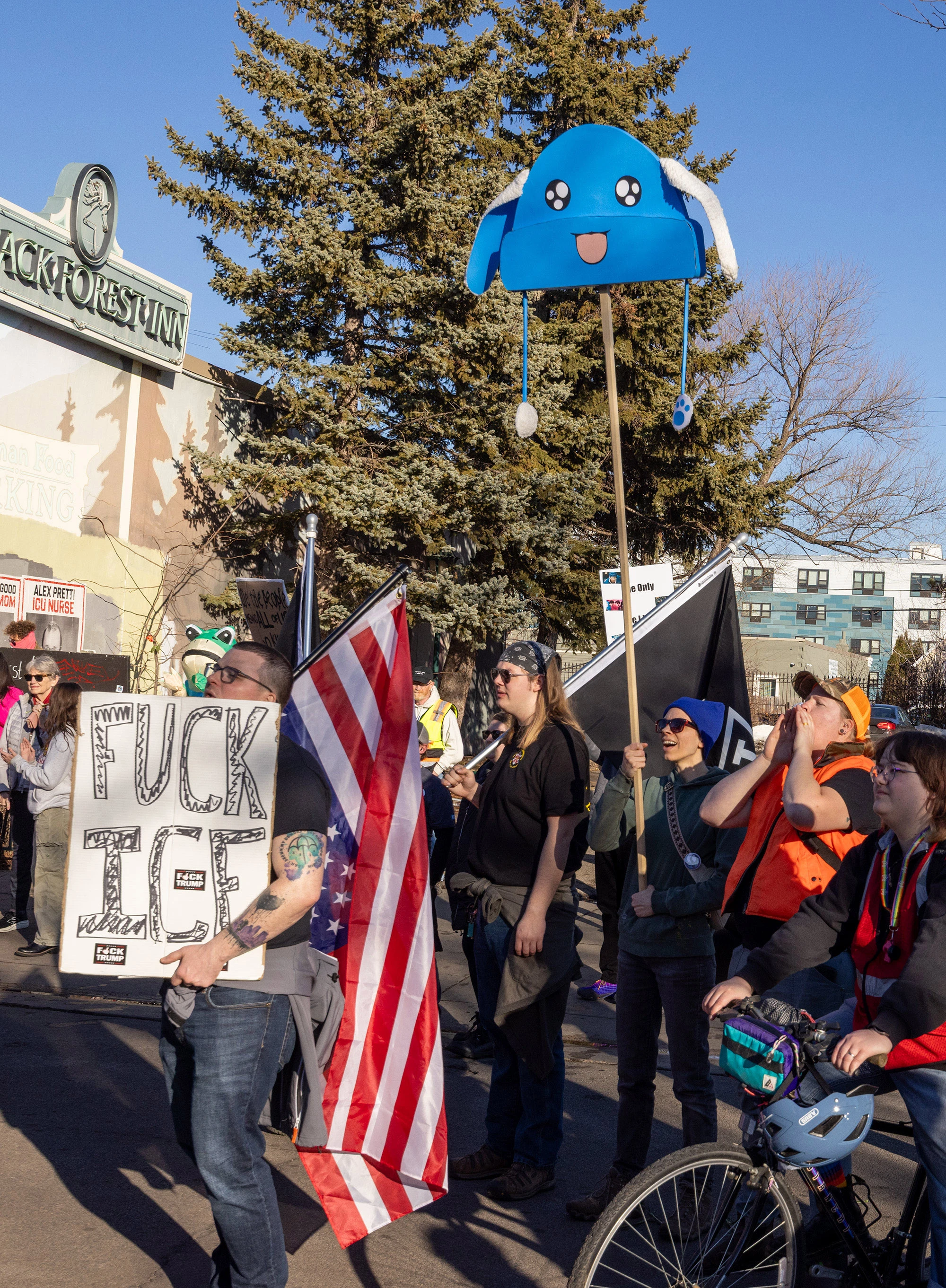 A tall placard topped with a large blue cartoon-style hat held high above a crowd of demonstrators at a Minneapolis anti-ICE protest, with people holding flags and other signs in the background.