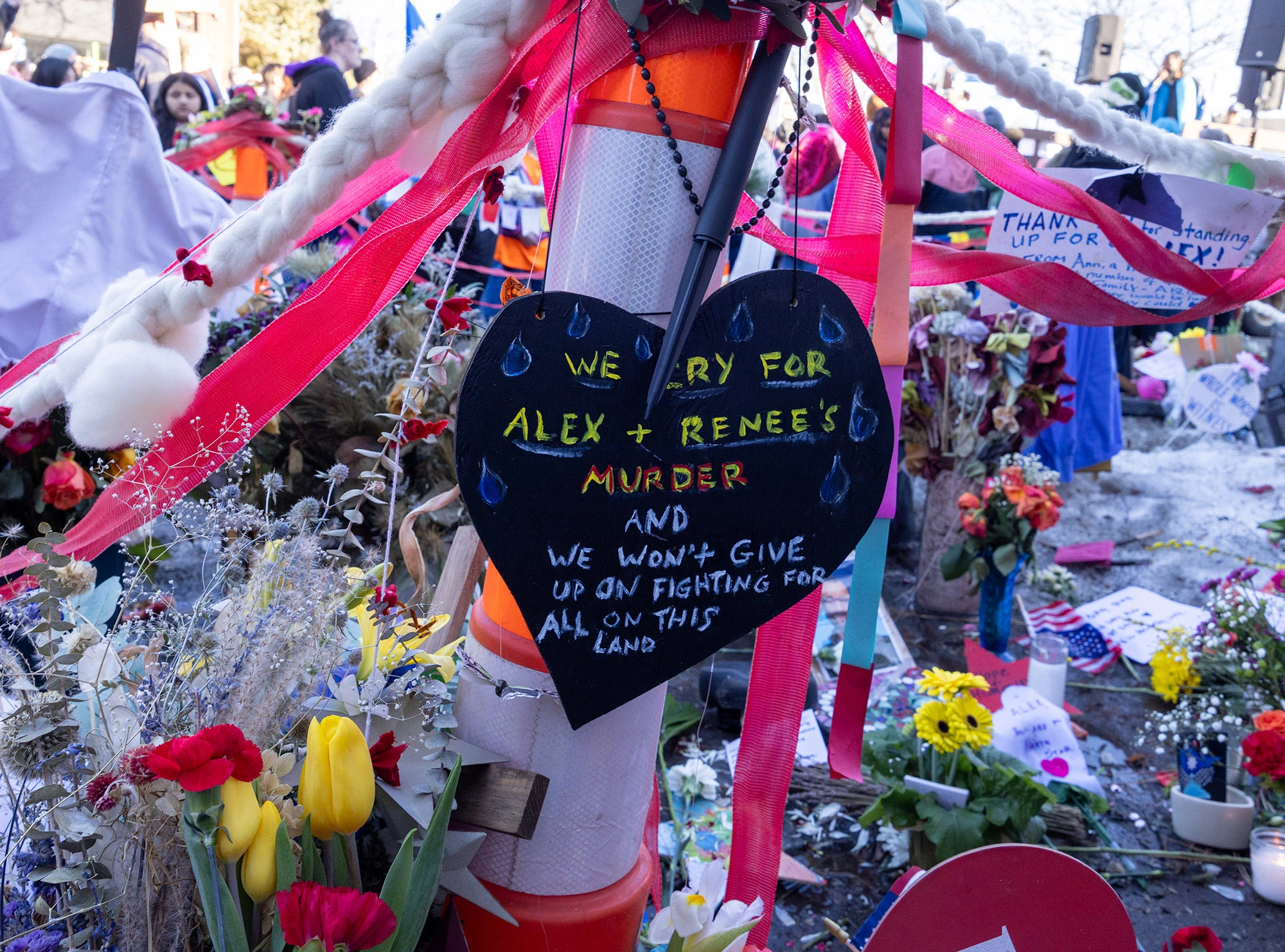 A close-up of a black heart-shaped sign hanging among ribbons, flowers and other memorial items at the Alex Pretti and Renée Good memorial in Minneapolis, with text expressing sorrow for their deaths and a commitment to keep fighting.