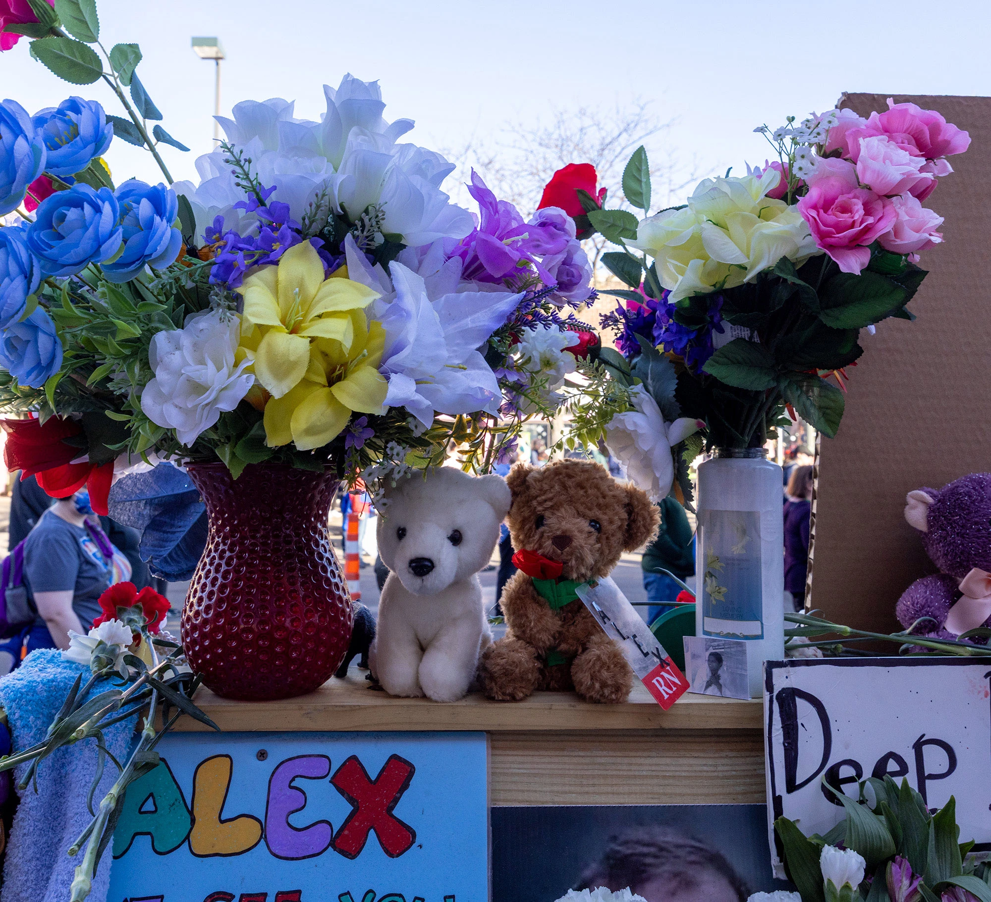 Two small teddy bears nestled among bouquets of flowers at the Alex Pretti memorial in Minneapolis, symbolizing tenderness and remembrance amid the larger collection of floral tributes.