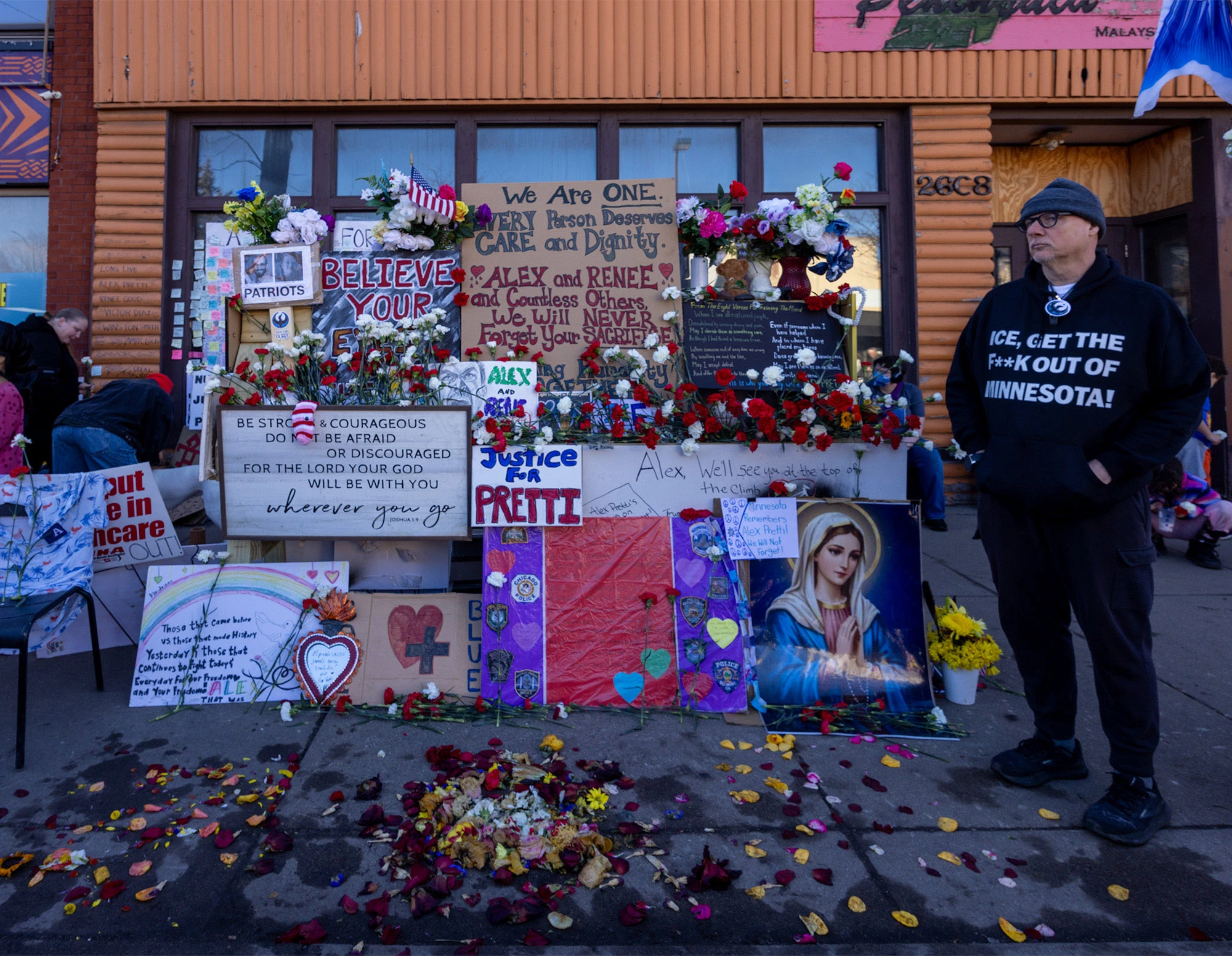 A wall of flowers, handmade signs, and messages honoring Alex Pretti and Renée Good at a makeshift memorial in Minneapolis, with flowers scattered on the sidewalk and a supporter wearing an 'ICE, Get the F**k Out of Minnesota!' sweatshirt standing nearby.