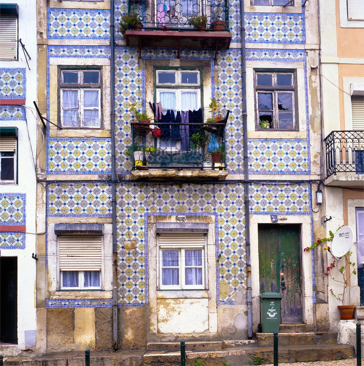 A tall Alfama facade clad in blue-and-yellow azulejo tiles, with iron balconies, laundry and potted plants, Lisbon