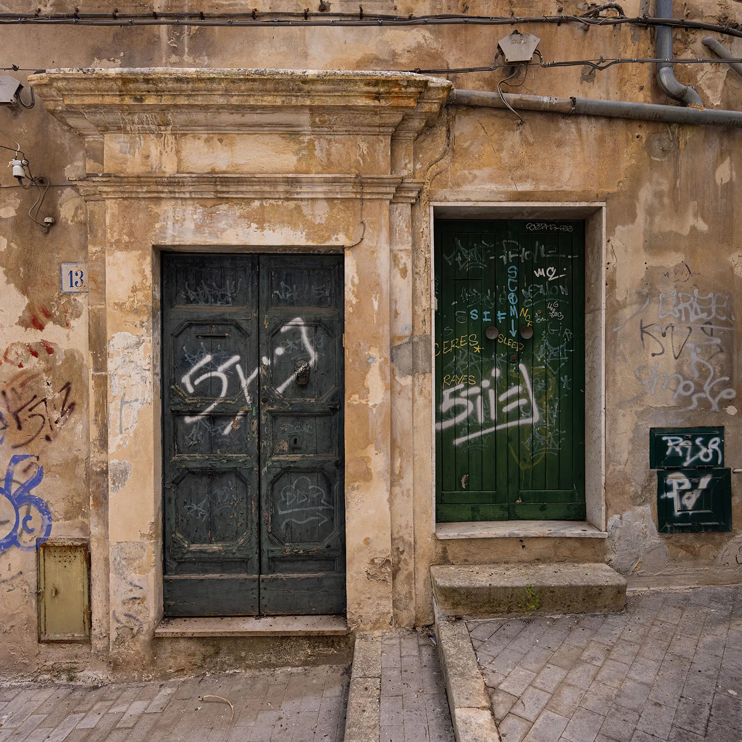Black and green doors marked with graffiti in a crumbling limestone alley in Modica, Sicily.