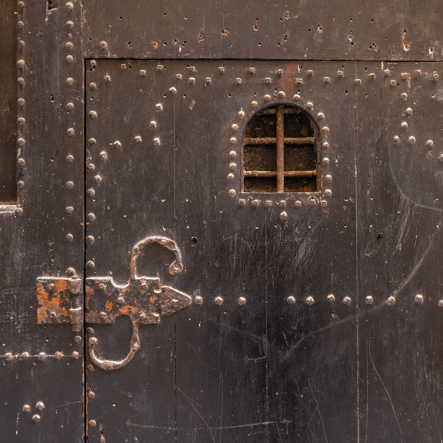 Old black door with iron studs, a small grilled peephole and a scrolled iron hinge in Palermo, Sicily.