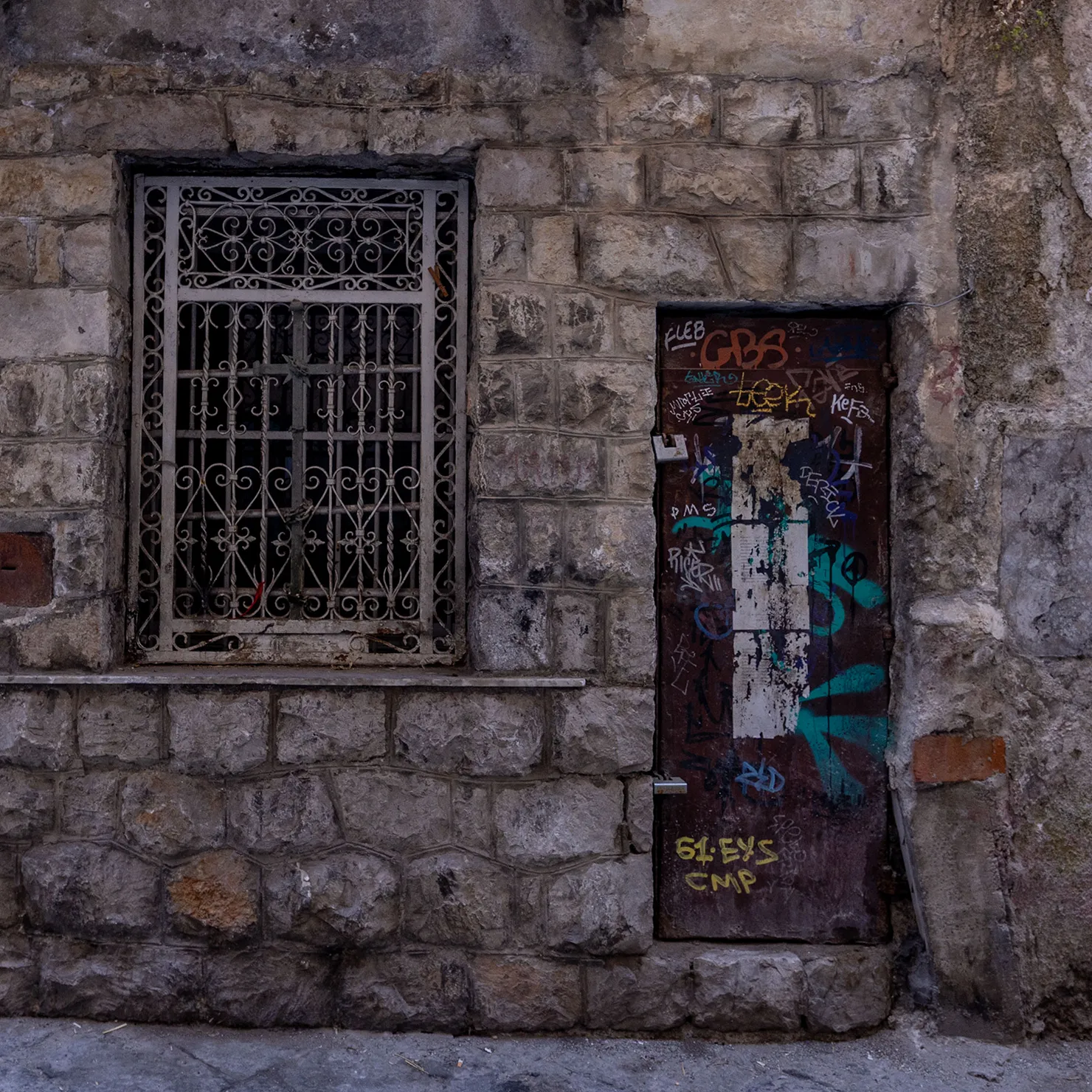 Narrow graffiti-covered door beside an iron-grilled window in a rough stone wall in Palermo, Sicily.