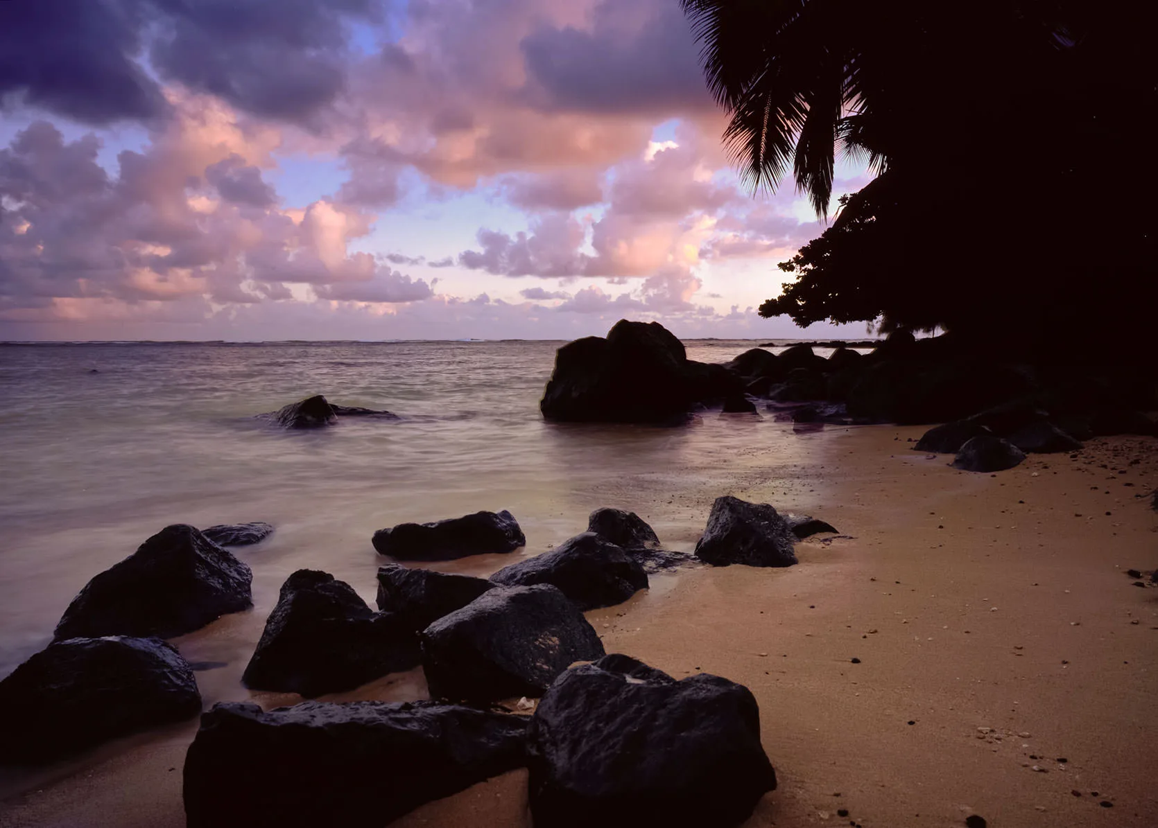 Dark lava rocks on the sand at Anini Beach, Kauai, under a purple and pink sunset sky.