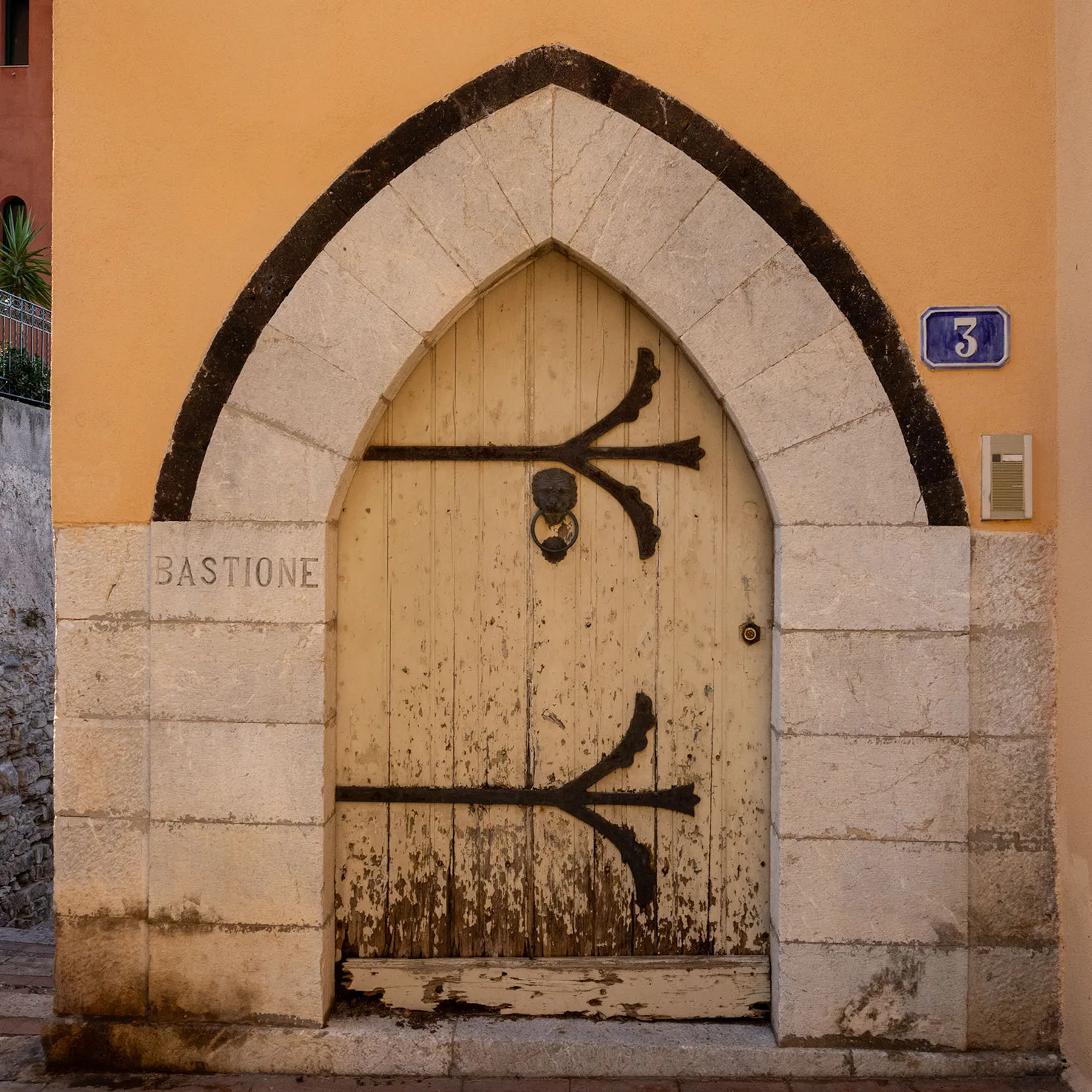 Weathered wooden door with black iron strap hinges under a pointed stone arch marked Bastione in Taormina, Sicily.