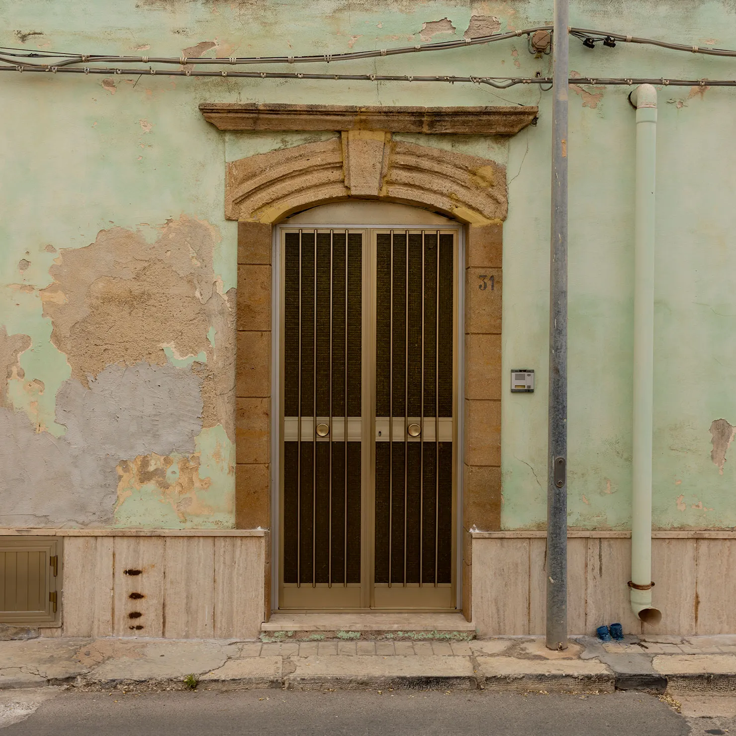 Bronze grille double door beneath a carved stone lintel on a peeling mint-green wall in Marzamemi, Sicily.