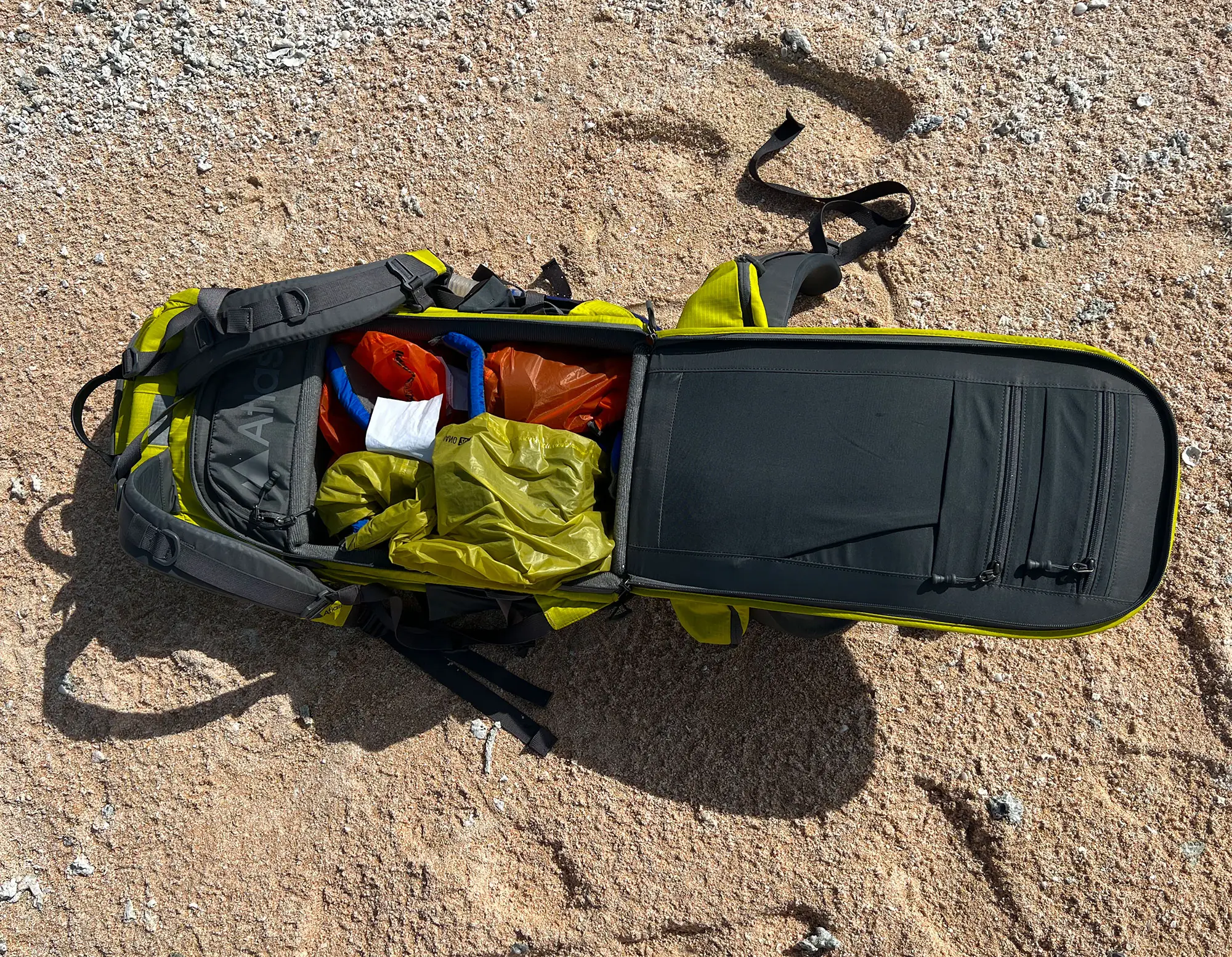 An open Atlas backpack on sand with camera lenses packed in colorful dry bags.