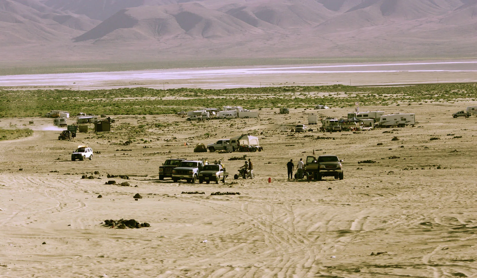 An informal desert camp of RVs, trucks and ATVs scattered across a sandy Nevada flat below barren mountains and a distant playa