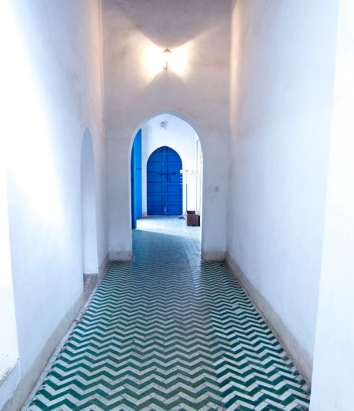 Sunlit white corridor with green zigzag tiles leading to a blue arched door in Marrakech's Bahia Palace.