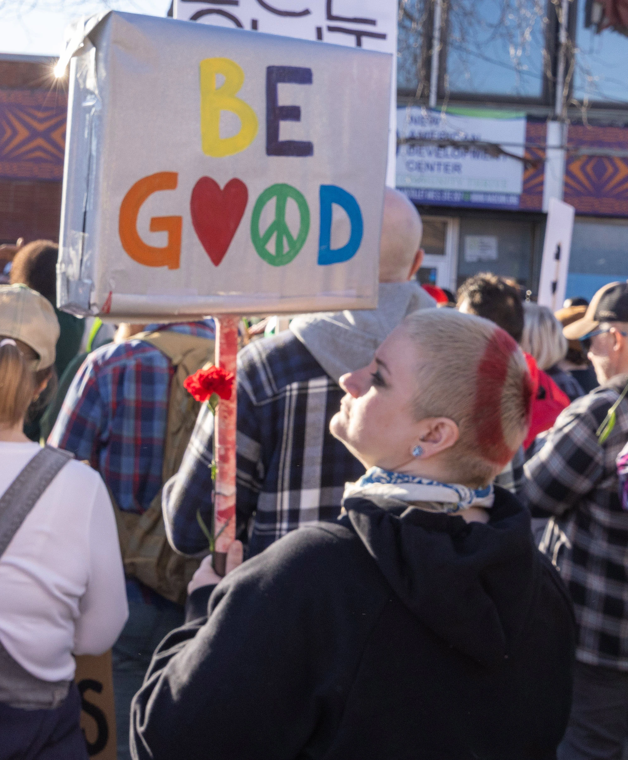 A protester seen from behind holding a sign reading 'Be Good' above a crowd at the Alex Pretti memorial and protest in Minneapolis, capturing a moment of peaceful expression among the demonstrators.