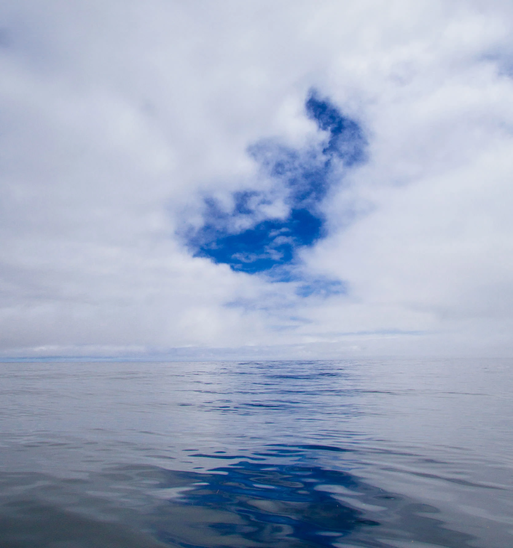 Open Pacific Ocean under a low marine cloud deck, with a break in the clouds revealing a deep blue patch of sky above calm reflective water about sixty miles offshore from Newport, Oregon.