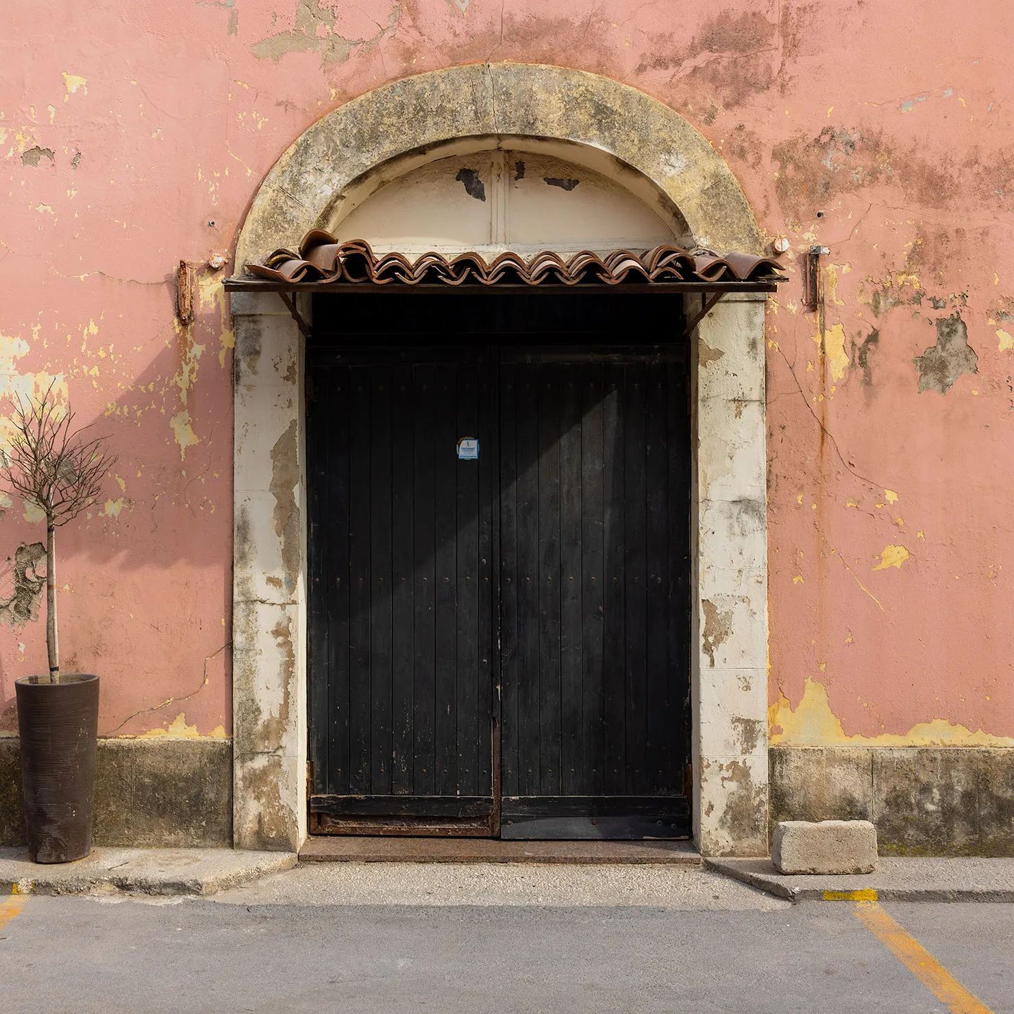 Black double door beneath a small tiled hood and stone arch on a rose-pink wall in Marzamemi, Sicily.
