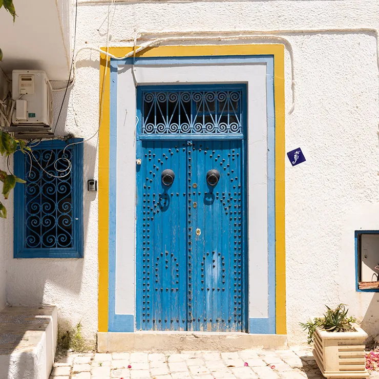 Blue wooden door with ornamental nail patterns and matching window grille, framed by yellow and blue trim in Sidi Bou Said, Tunisia.