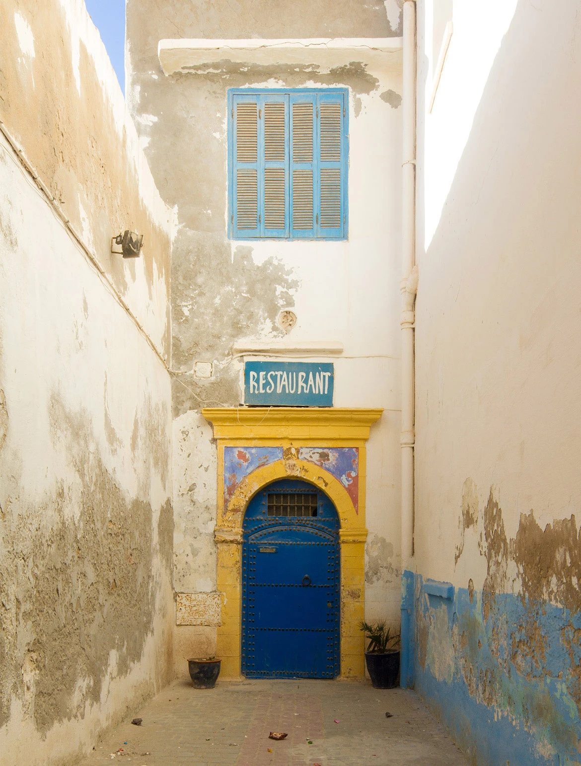Blue arched door beneath a yellow frame and shuttered window in a narrow Essaouira alley, Morocco