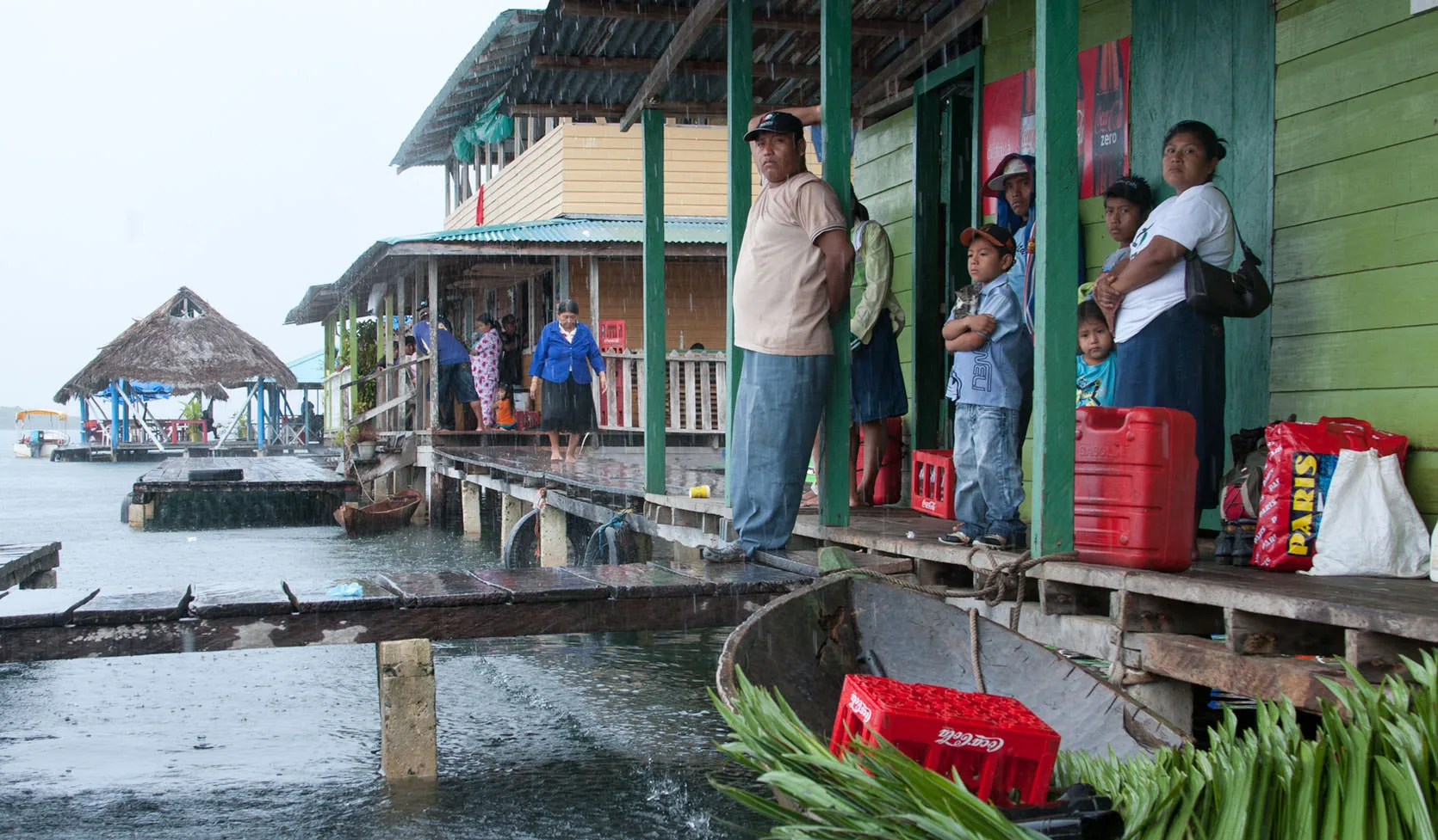 People sheltering from the rain on the porch of a green over-water shop stacked with soda crates and fuel jugs, Bocas del Toro