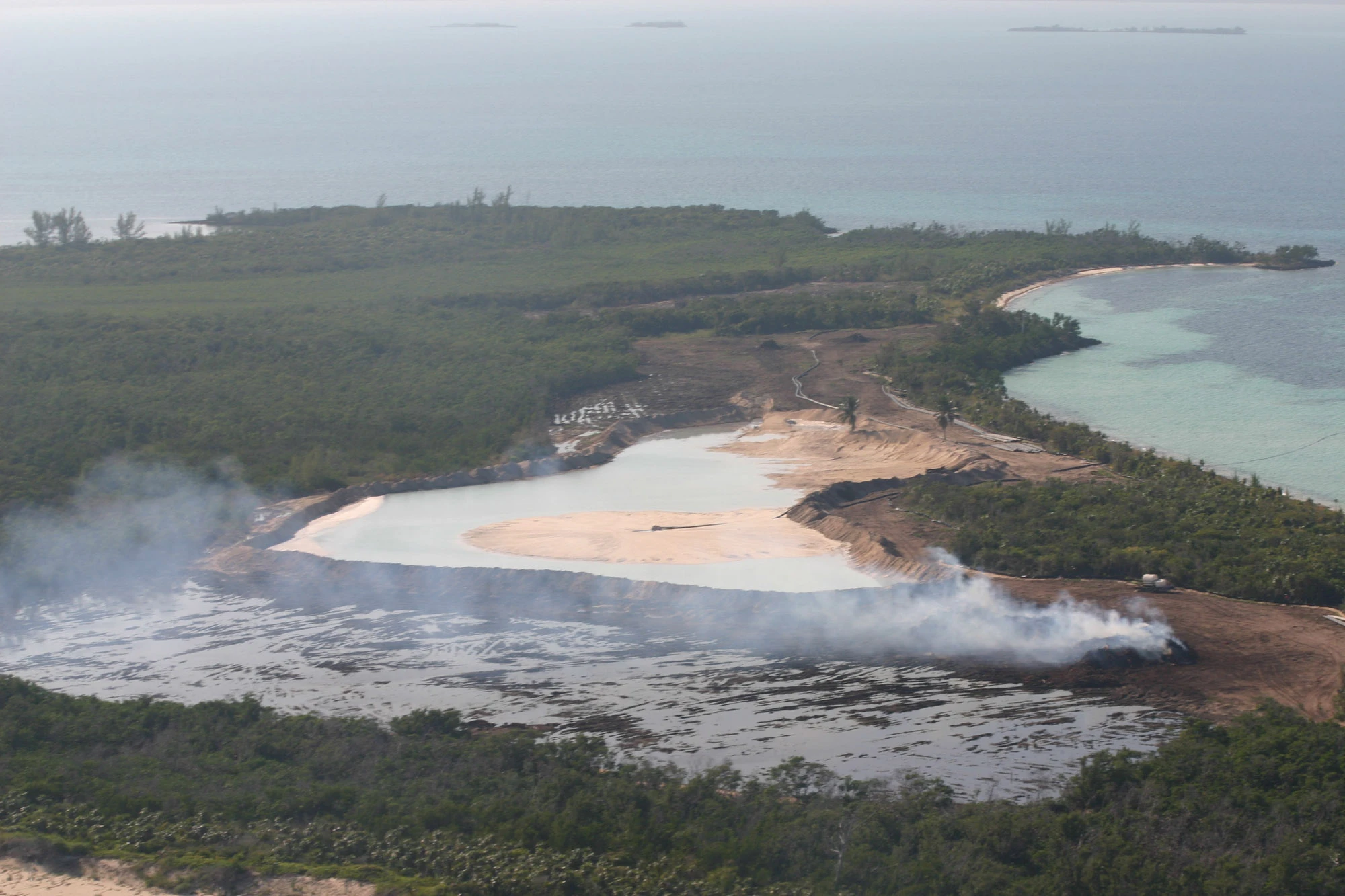 An aerial photograph showing mangroves burning at Baker's Bay while marina construction continues in the background, part of the pattern of habitat loss documented across the eleven site-visit reports tabulated below.