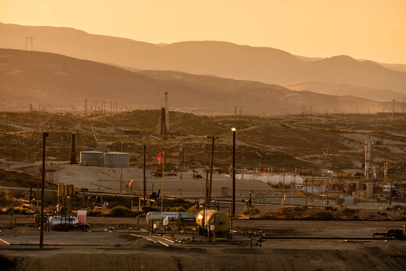 Oil wells, tanks and pipelines near Buttonwillow in California's Central Valley, under hazy golden light with hills beyond.