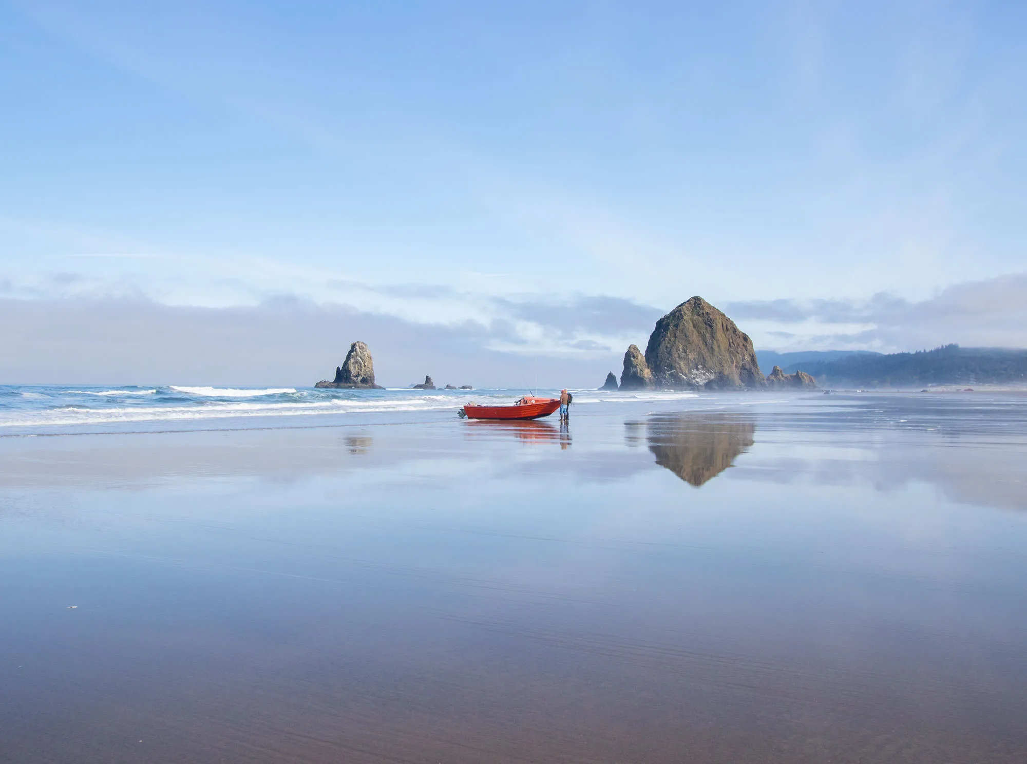 A red beach-launched fishing dory on the wet sand at Cannon Beach, with Haystack Rock reflected in the shallows.