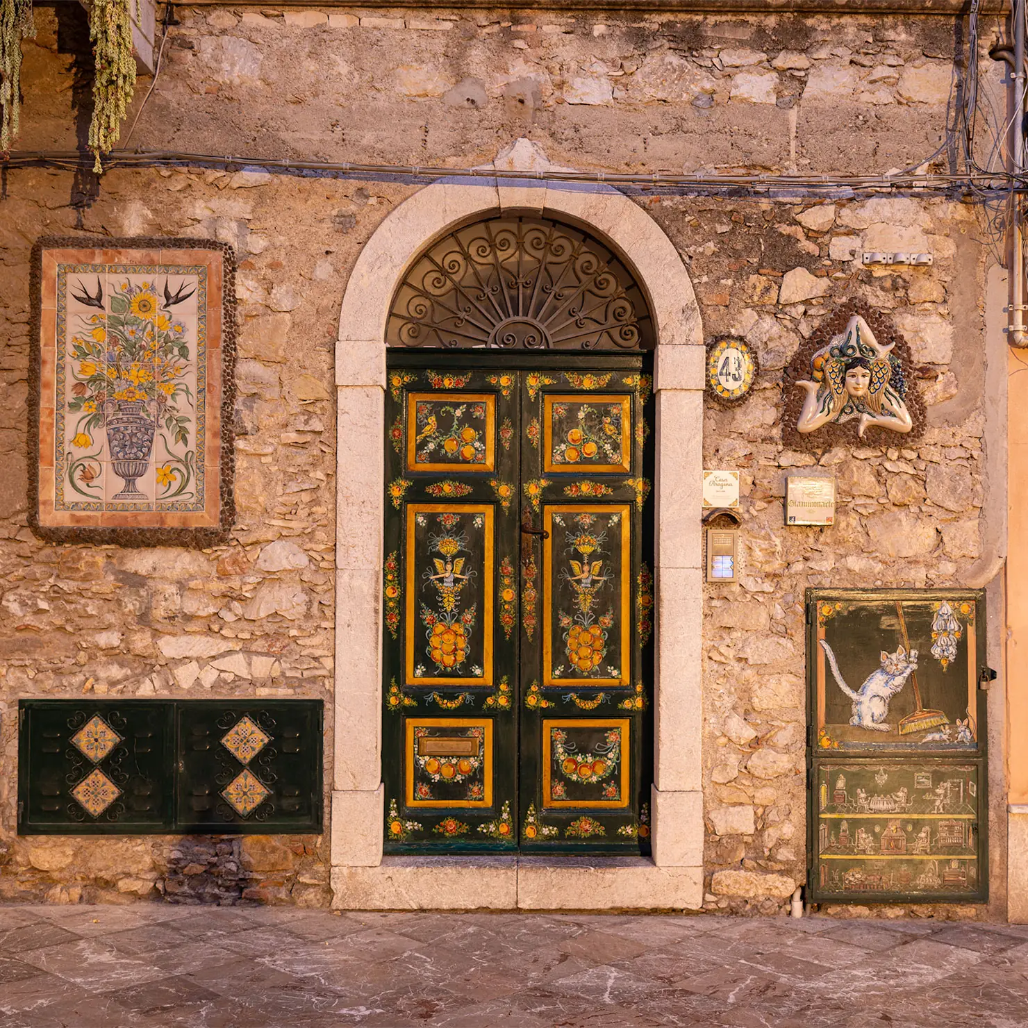 Dark green arched door hand-painted with oranges and flowers, framed by painted tiles and a Trinacria emblem in Taormina, Sicily.