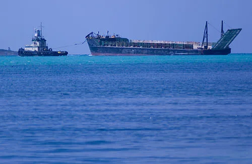 A tugboat towing a construction barge across the turquoise shallows off Great Guana Cay