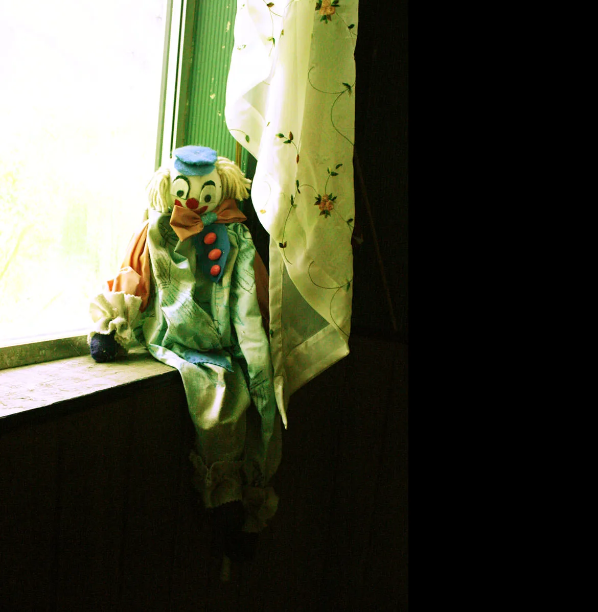 A faded clown doll sitting on a sunlit windowsill beside a curtain in a workshop in Austin, Nevada.
