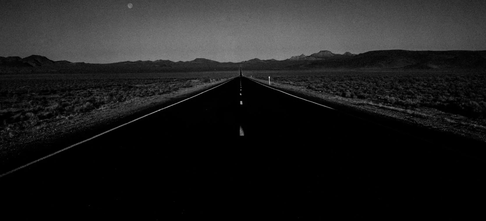 A dark, empty desert highway running arrow-straight toward distant mountains under a rising moon on the Extraterrestrial Highway near Rachel, Nevada
