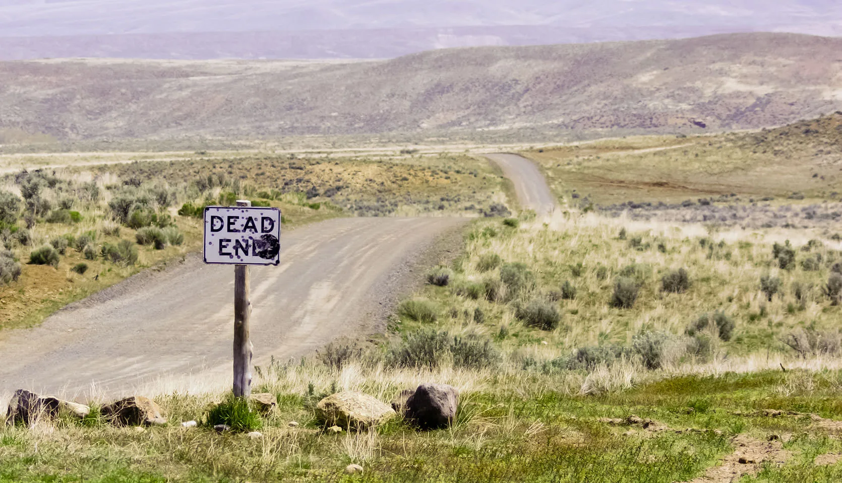 A weathered 'Dead End' sign where a lonely gravel road winds off through the sagebrush high desert of the Owyhee country