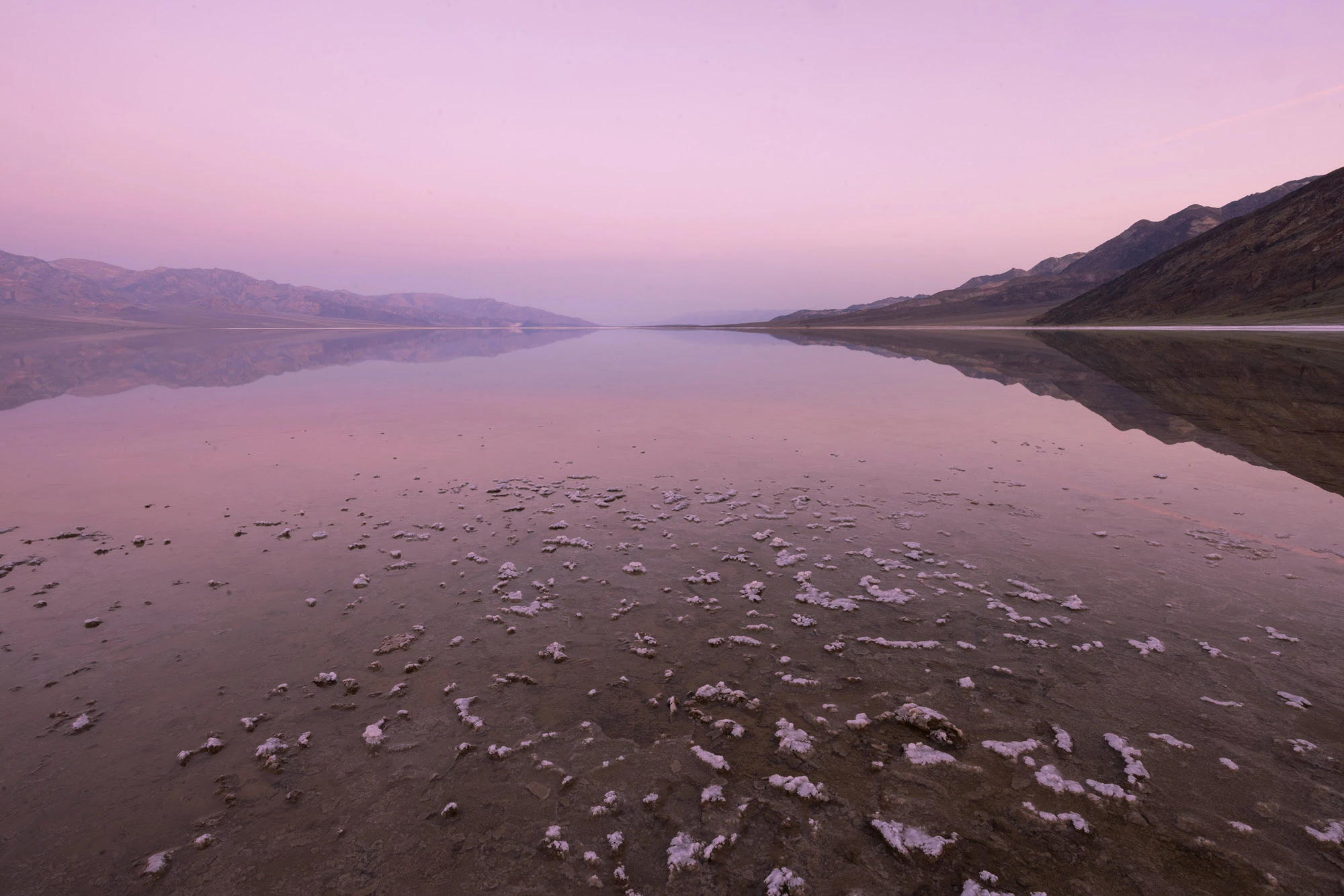 A calm, mirror-like expanse of Lake Manly reflects pastel pink and lavender skies in Death Valley, with scattered salt formations in the foreground and mountain ranges fading into the distance.