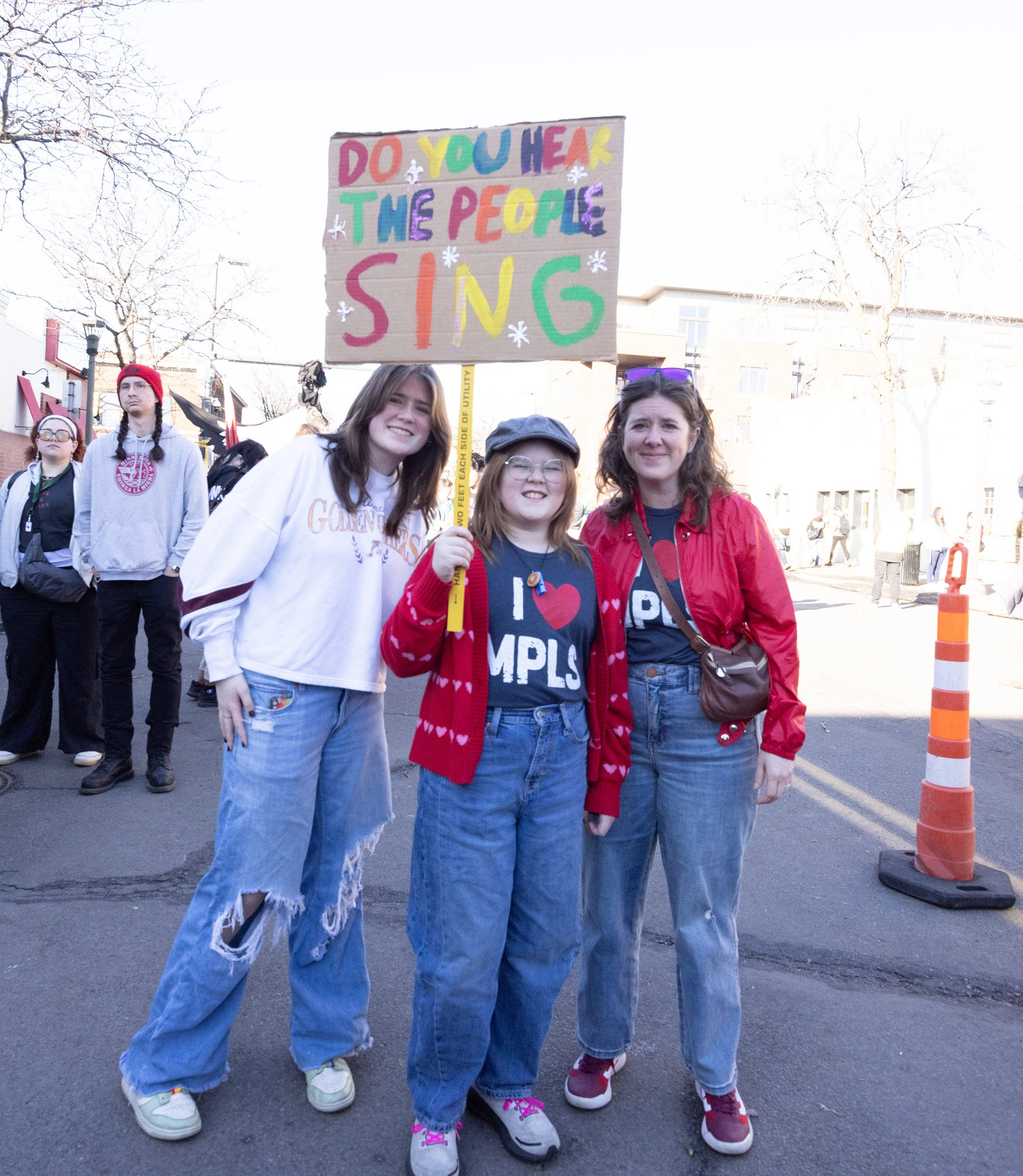 A mother and her two daughters standing together in the crowd at the Alex Pretti memorial and protest in Minneapolis, one holding a colorful sign above their heads.