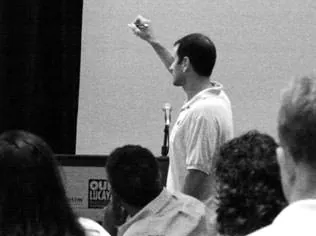 Black-and-white photo of a man pointing at a projected screen while speaking to an audience.
