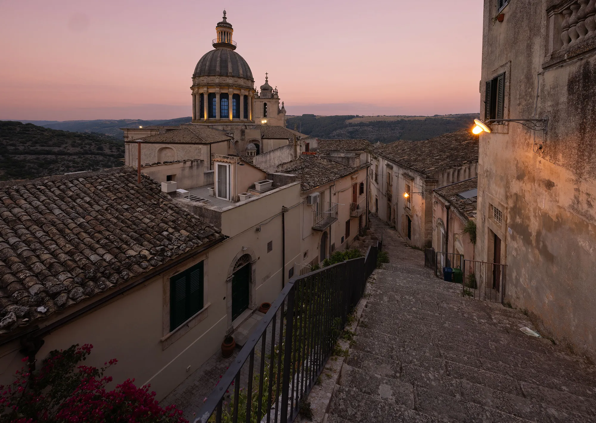 The dome of the Duomo di San Giorgio rising over the rooftops of Ragusa Ibla at dusk, with a stone stairway descending beside it.