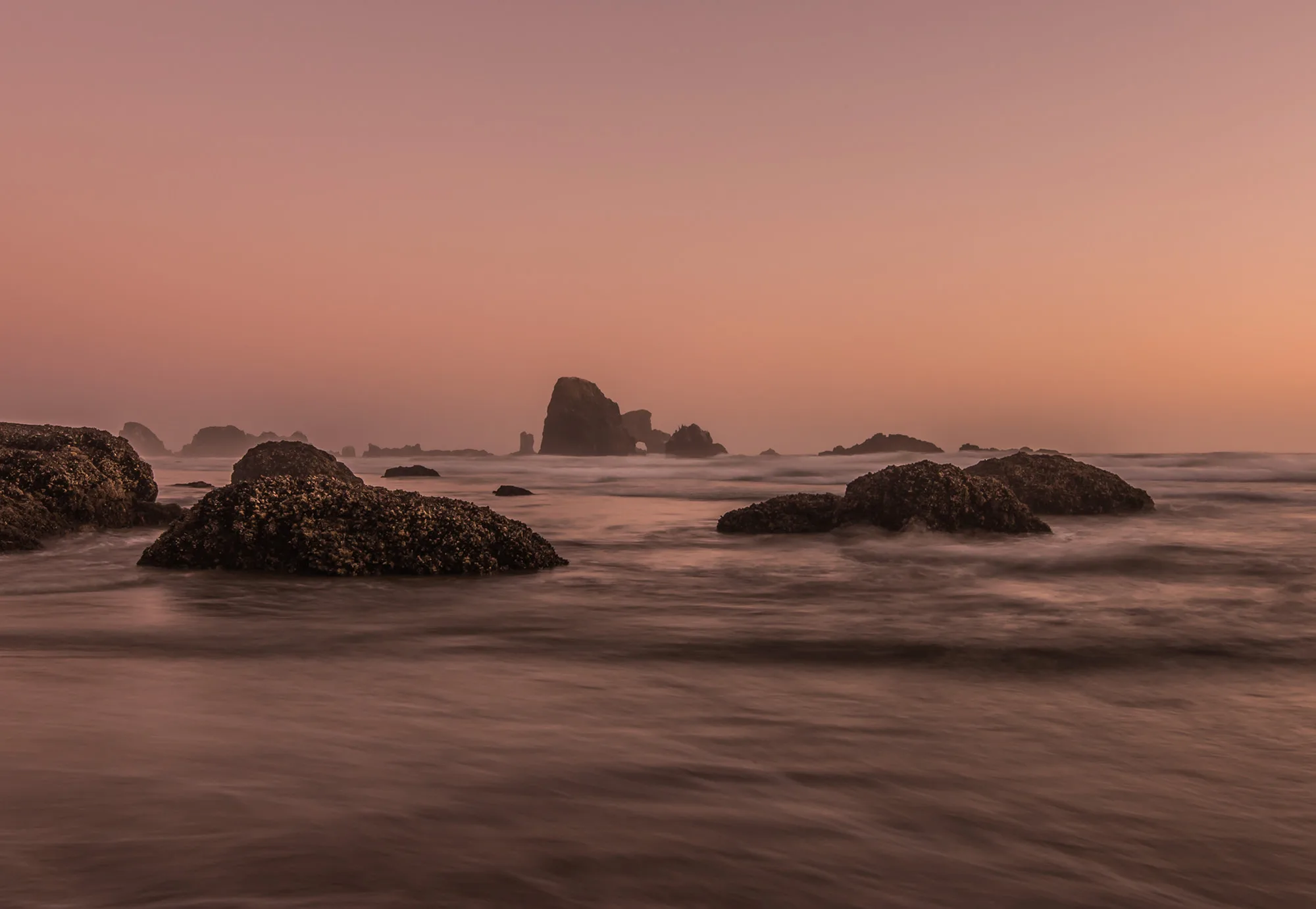 Submarine Rock and smaller sea stacks seen from the south end of Indian Beach at pink dusk, waves blurred to mist.