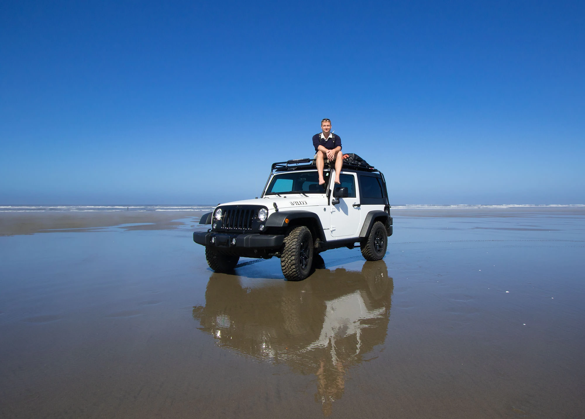 Erik Gauger sits on the roof of a white Jeep Wrangler parked on a wet, reflective beach along the Oregon Coast. The ocean and horizon are visible in the background under a clear blue sky.