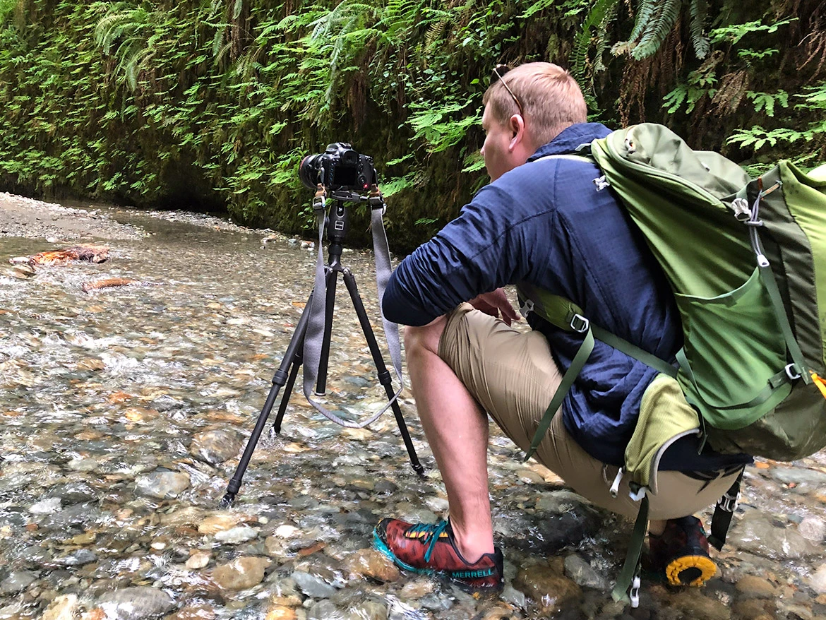 Erik Gauger crouches in a shallow riverbed in California with a camera mounted on a tripod, wearing a green backpack and water shoes. Fern-covered canyon walls line the background.