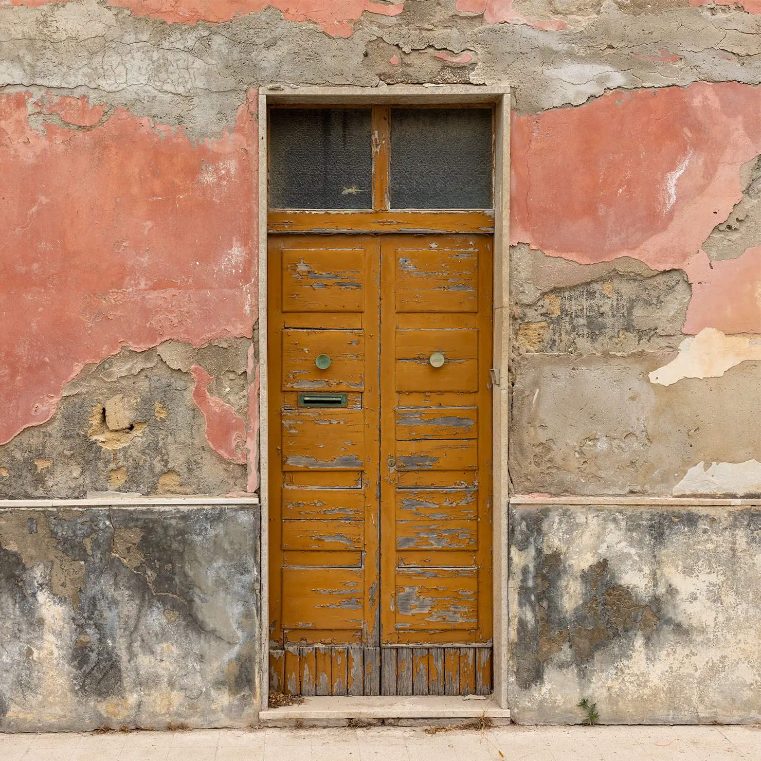 Worn orange wooden door with a transom window in a wall of peeling pink plaster in Marzamemi, Sicily.