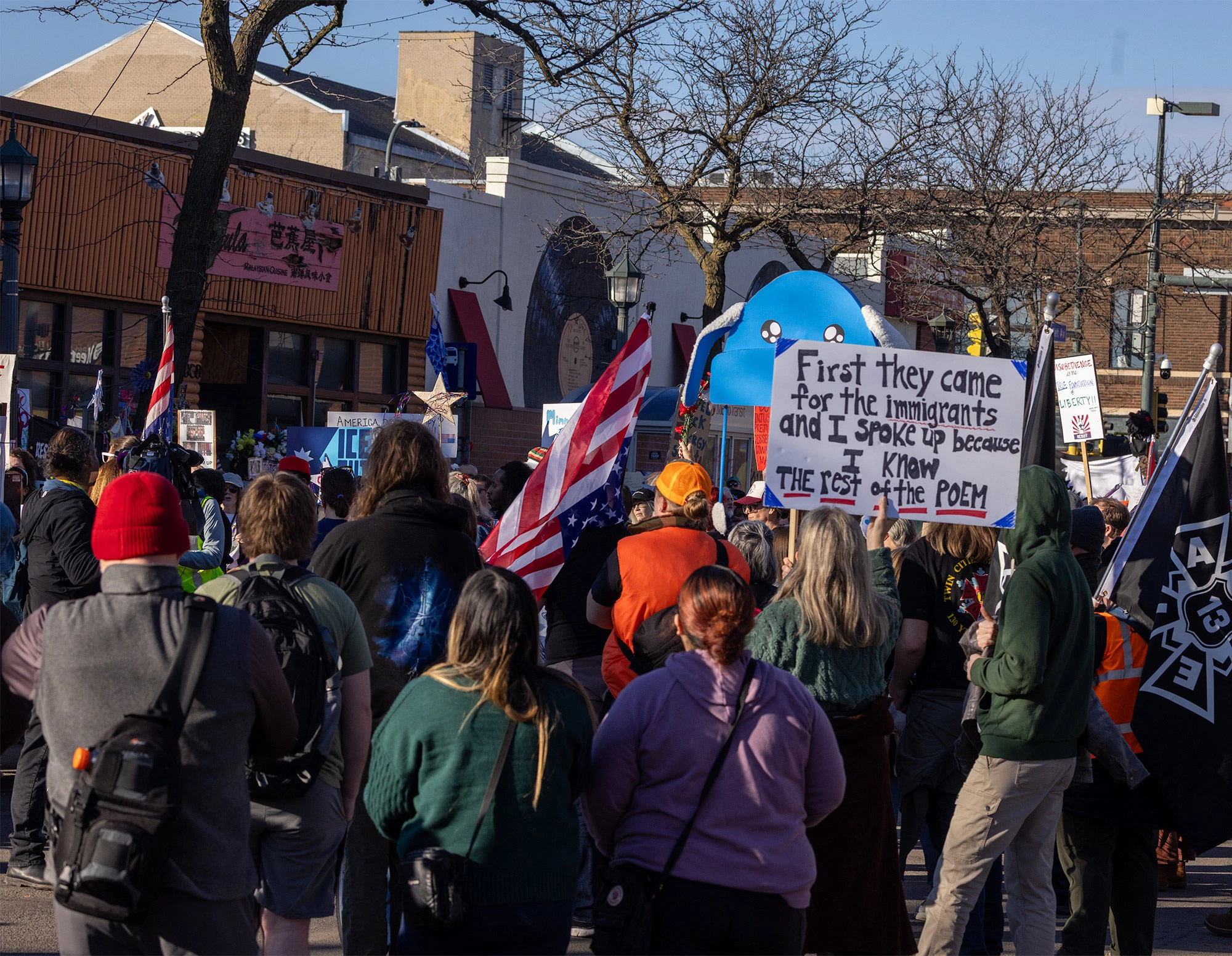 A large protest sign held above a crowd at the Alex Pretti vigil in Minneapolis, reading 'First they came for the immigrants and I spoke up because I knew the rest of the poem,' seen among other signs and memorial items in the street.