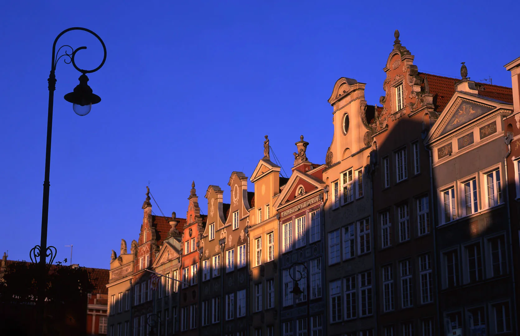 An ornate wrought-iron street lamp against a blue sky beside a row of gilded, gabled facades on Gdansk's Long Market