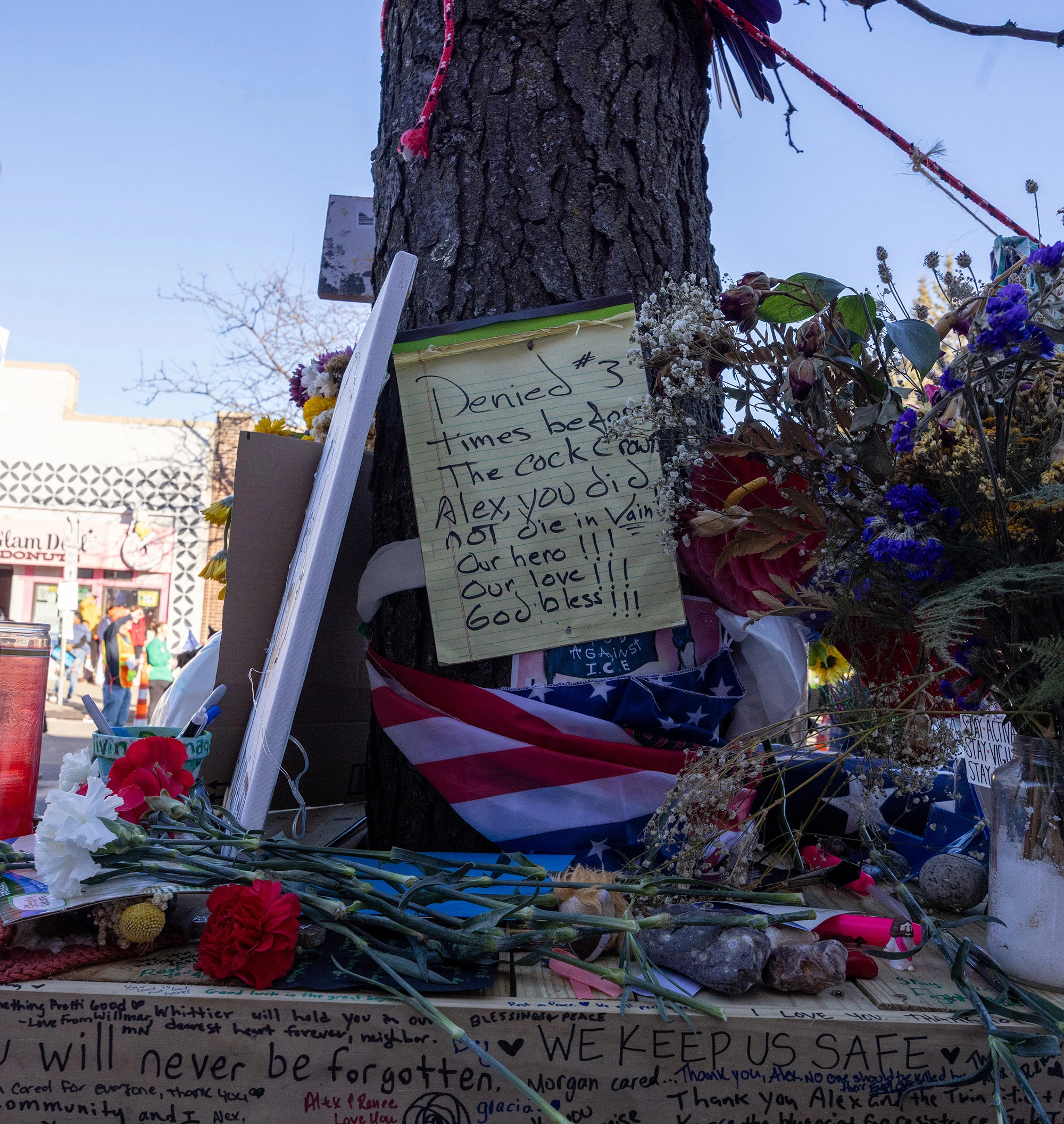 A handwritten memorial note and flowers placed at the Alex Pretti vigil in Minneapolis, attached to a tree with ribbons and surrounded by blossoms and other tributes.