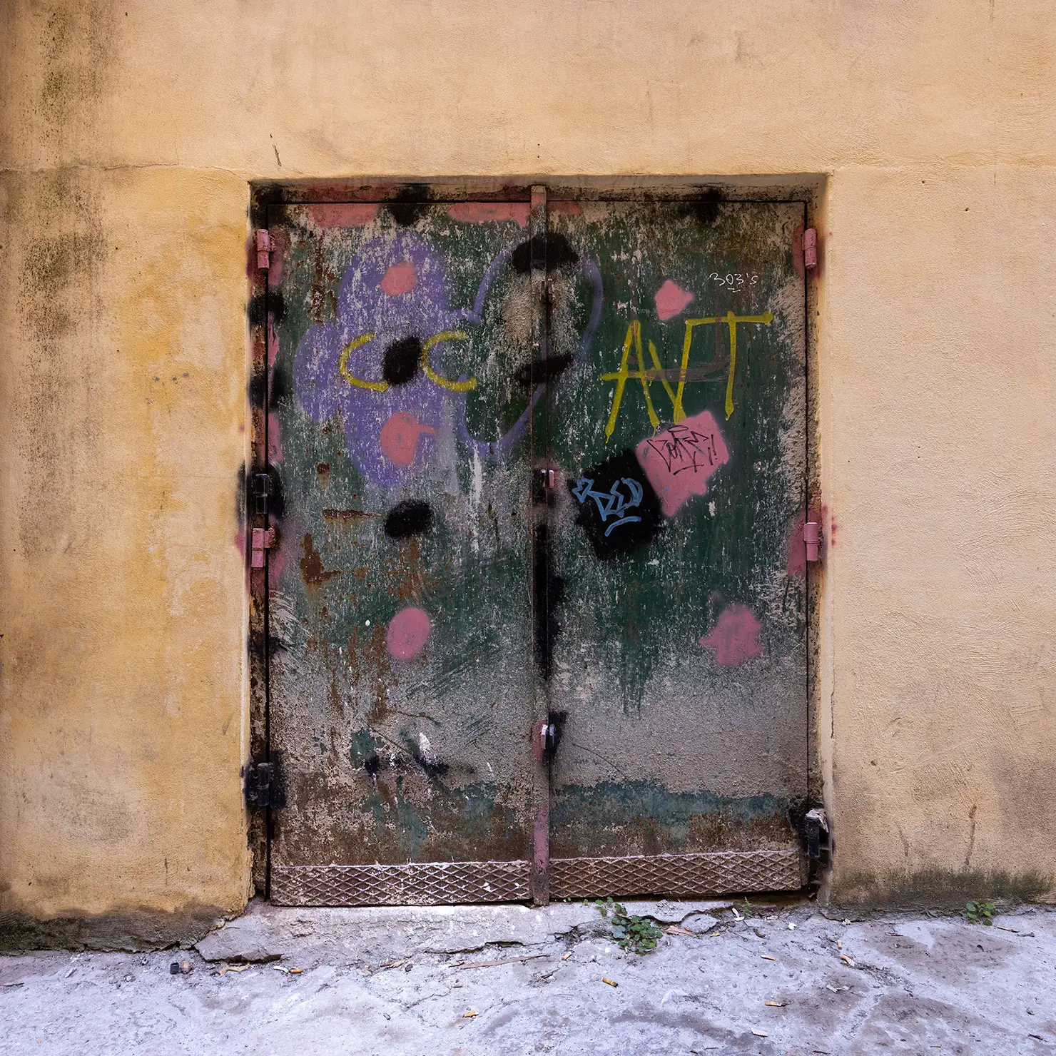 Green metal door covered in graffiti and torn posters in a pale ochre wall in Palermo, Sicily.