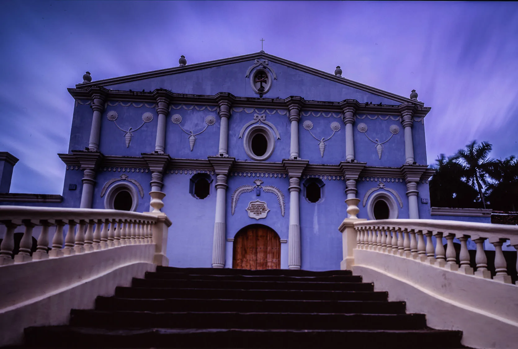 A pale blue neoclassical church facade with columns rising above a staircase at dusk, Granada
