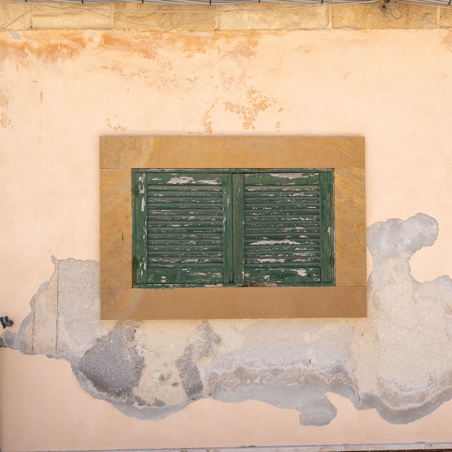Faded green window shutters in an ochre frame on a peeling peach plaster wall in Marzamemi, Sicily.