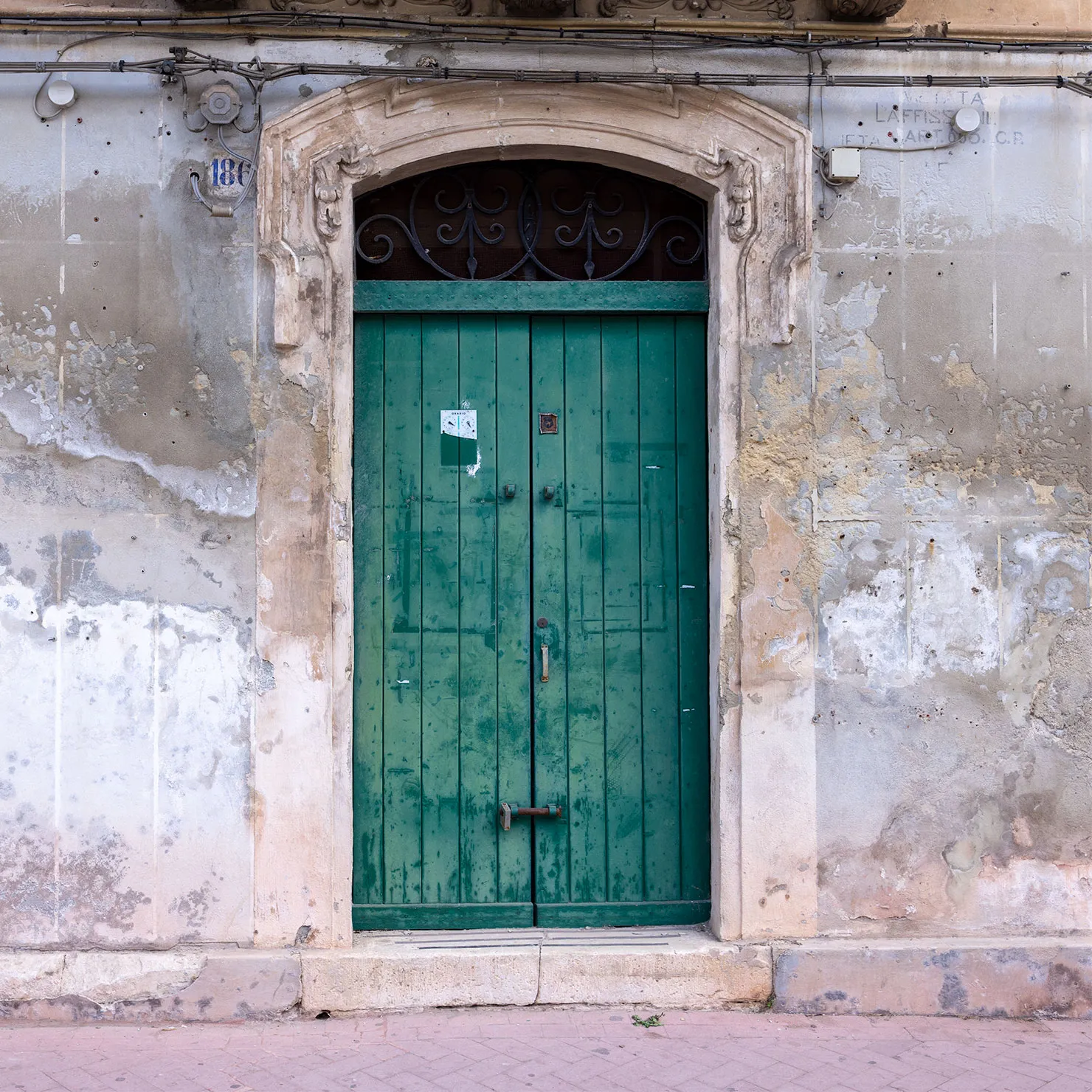 Deep green wooden double door beneath a carved stone lintel and iron fanlight on a peeling plaster wall in Modica, Sicily.