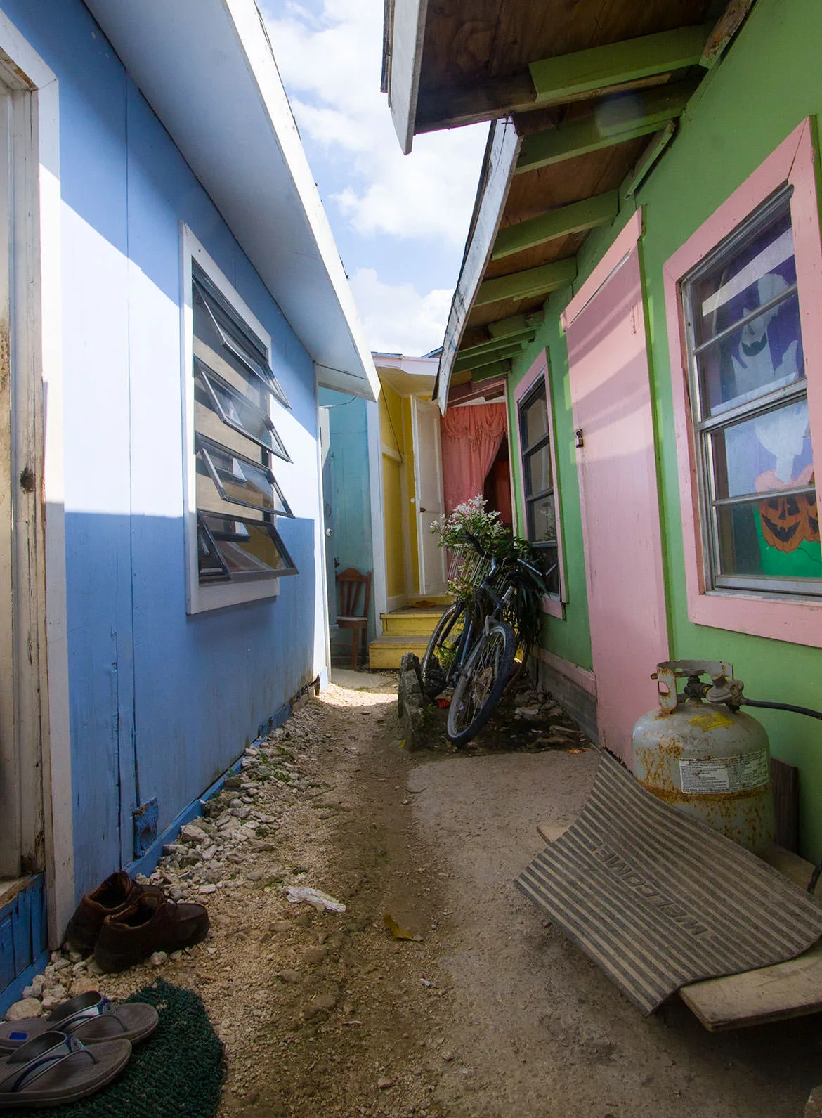 A narrow dirt alley between brightly painted plywood shacks with a bicycle and a welcome mat, a Haitian settlement near Treasure Cay, Abaco