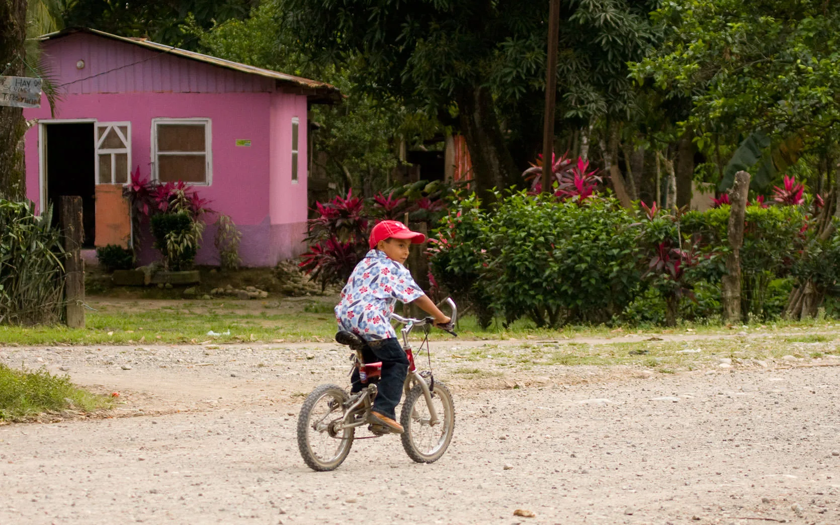 A boy in a red cap riding a bicycle on a dirt road past a pink house, Honduras