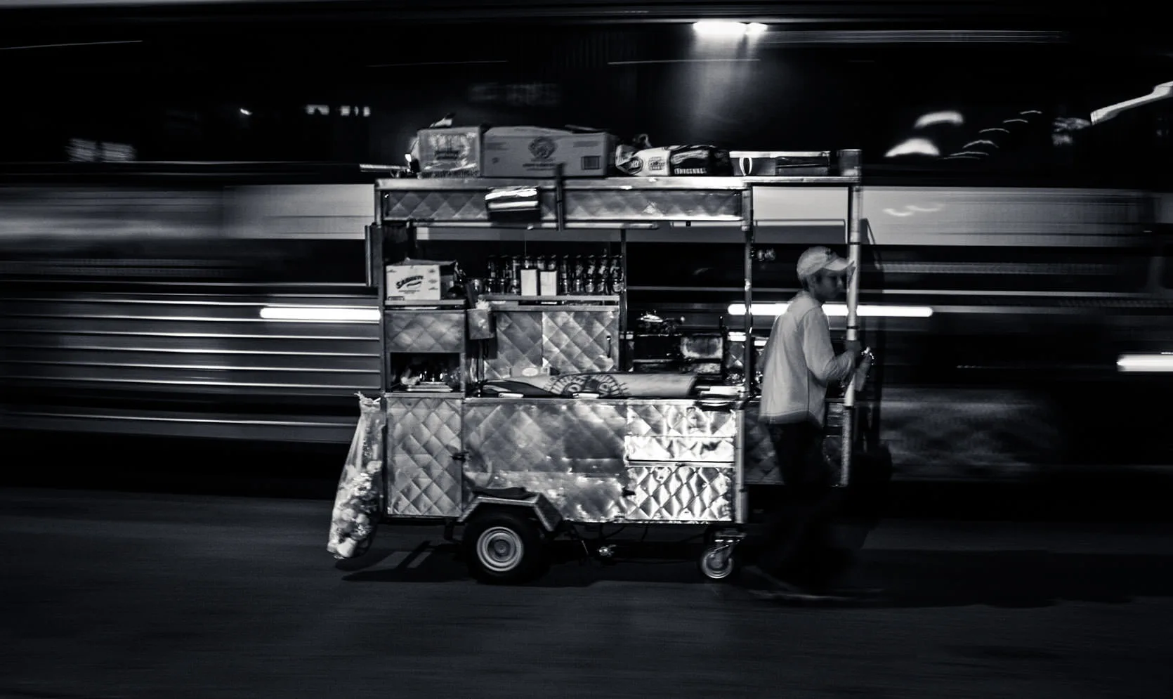 Black-and-white photo of a street vendor pulling his food cart past a blur of passing traffic at night in New York.