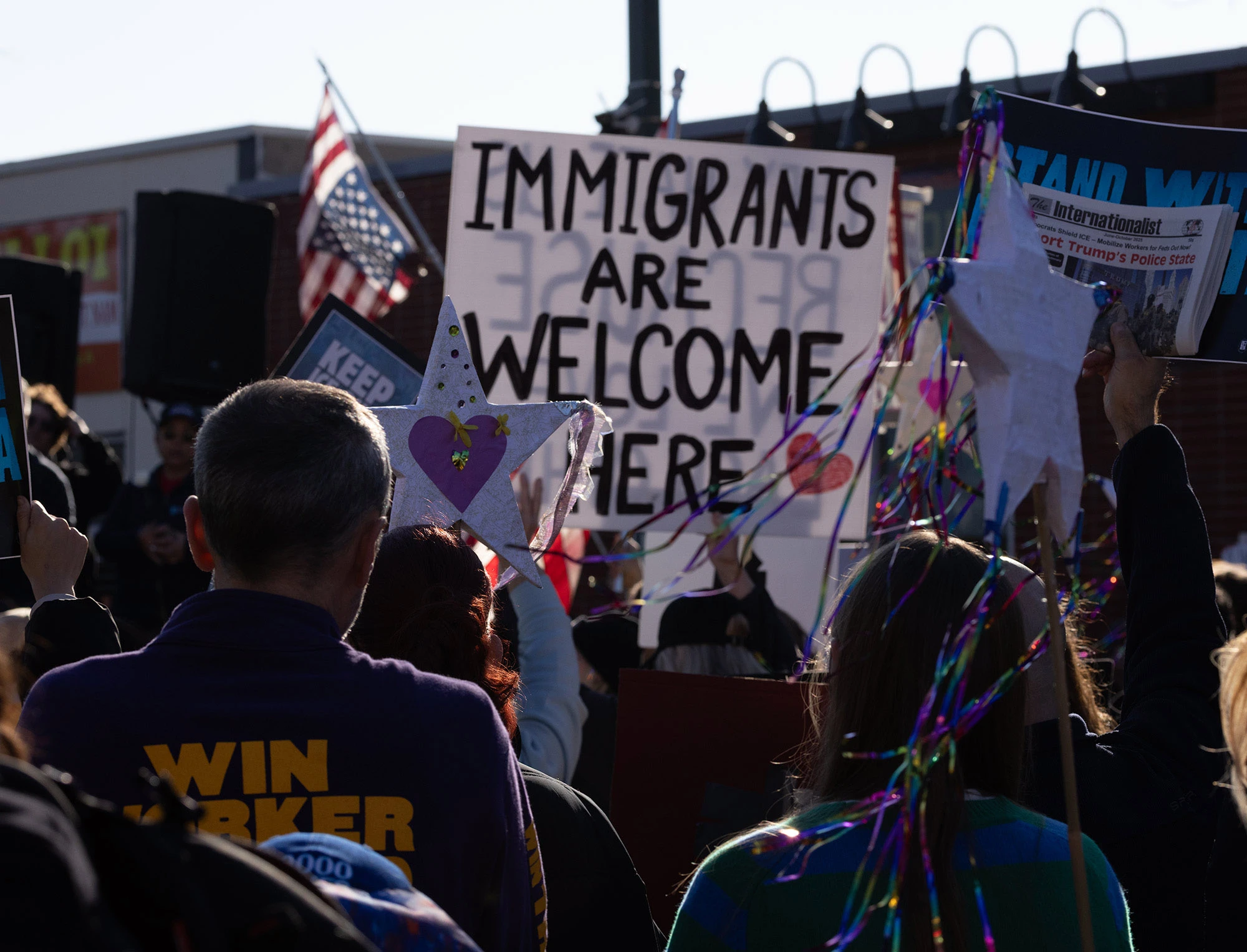 A crowd at the Alex Pretti memorial in Minneapolis holding a large handmade sign that says 'Immigrants are welcome here' among other protest signs.