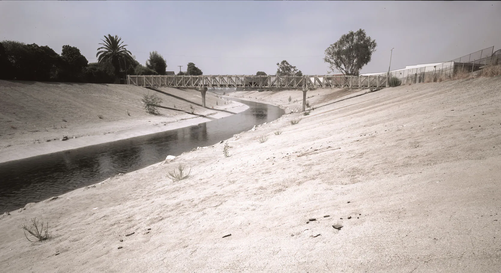 A wooden footbridge spanning the concrete channel of the Los Angeles River, a thin ribbon of water below, in black and white