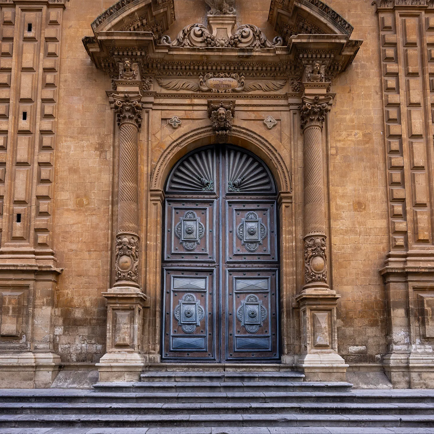 Carved blue-gray church door beneath an ornate Baroque stone portal and steps in Modica, Sicily.
