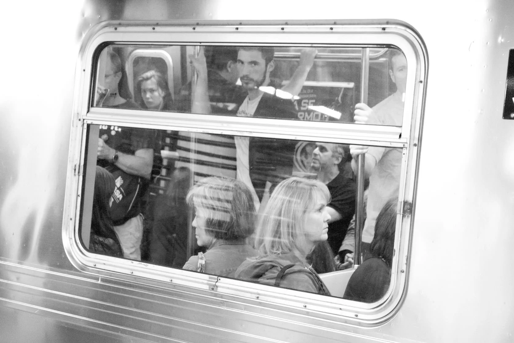 Black-and-white photo of passengers crowded inside a New York subway car, seen through its window.